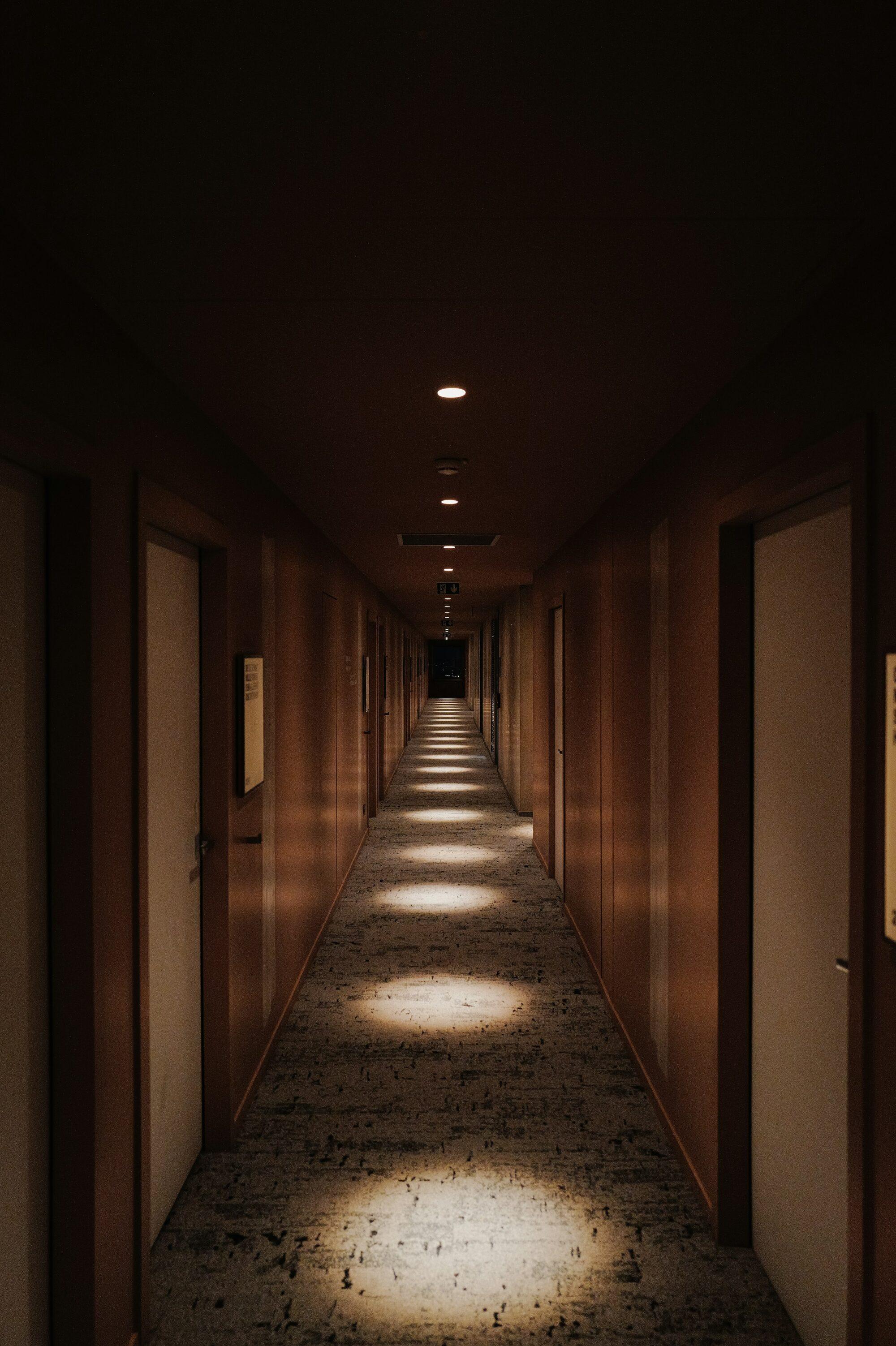 A quiet hotel corridor lined with dark timber panelling, ceiling downlights casting pools of light along the carpet