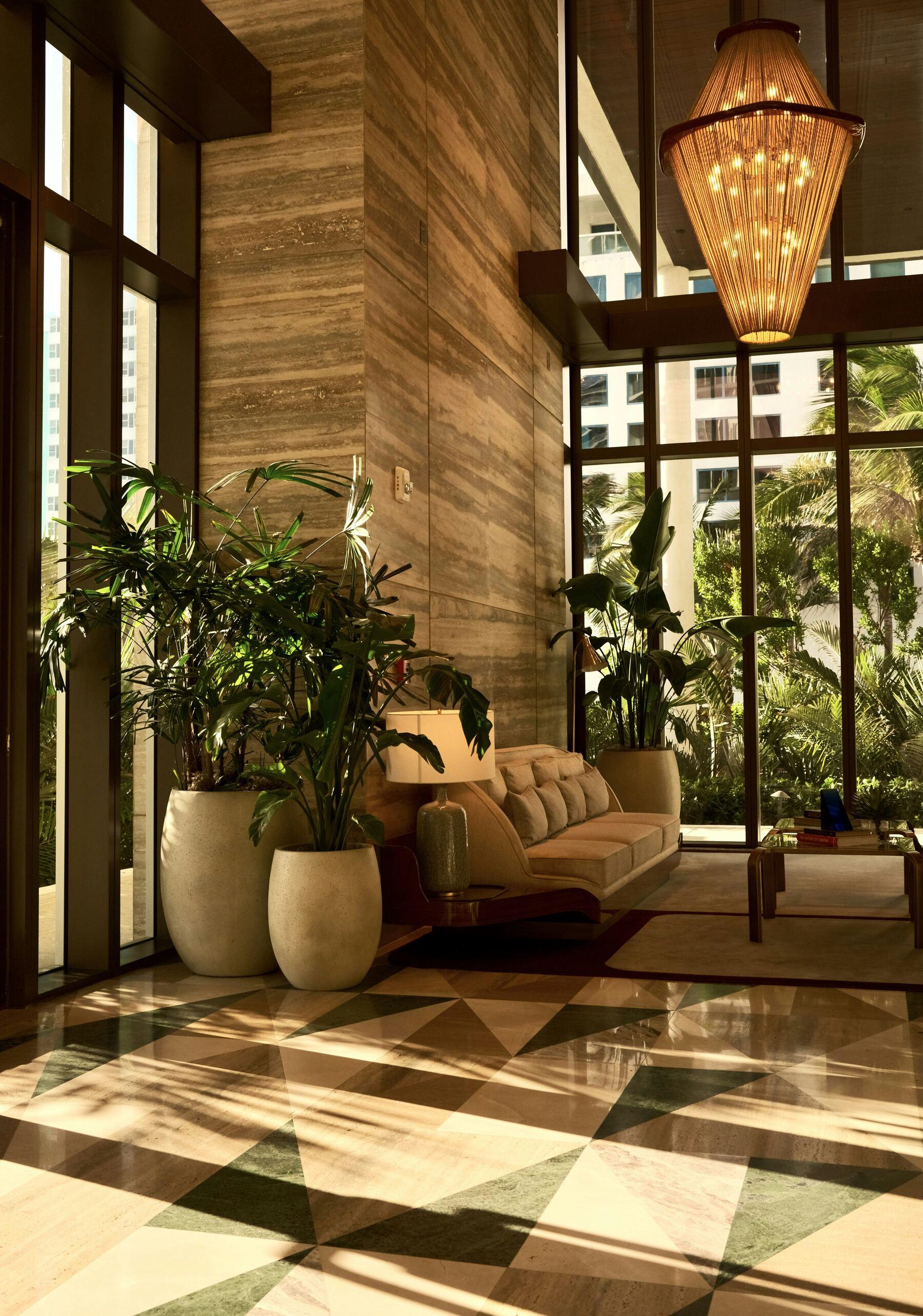 A sunlit hotel lobby with travertine walls, a woven pendant chandelier, potted palms and a marble floor laid in triangles